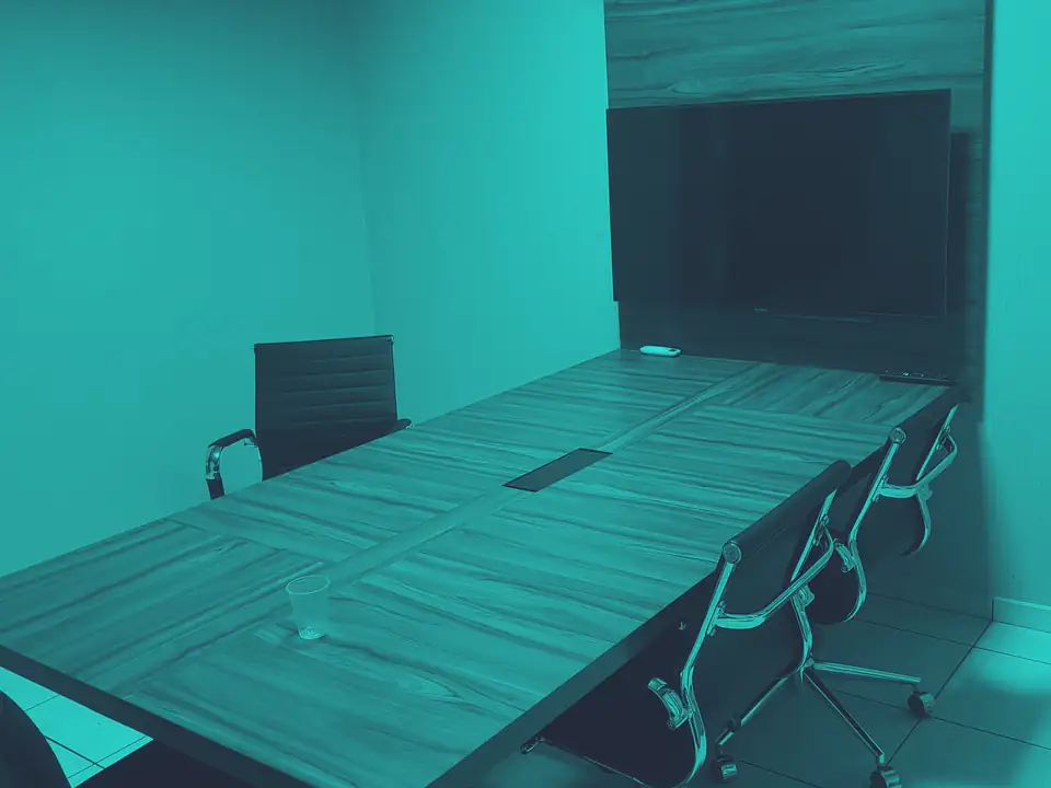 Sala de reunião da segunda sede, com mesa longa, cadeiras e uma televisão na parede