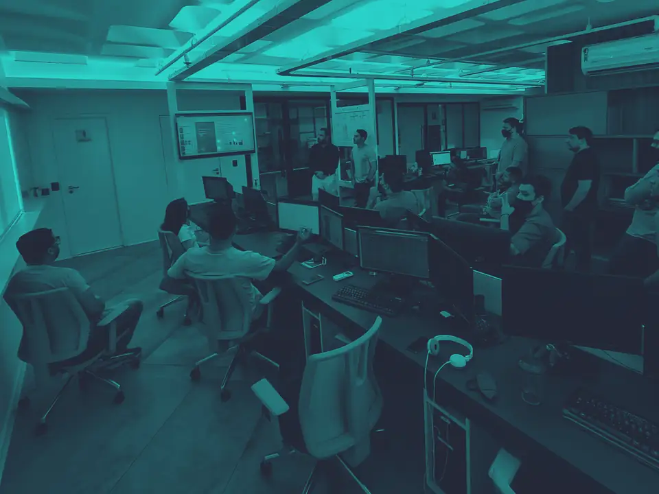 Time trabalhando no open space da sede atual, com estações de dois monitores e painéis de acompanhamento ao fundo